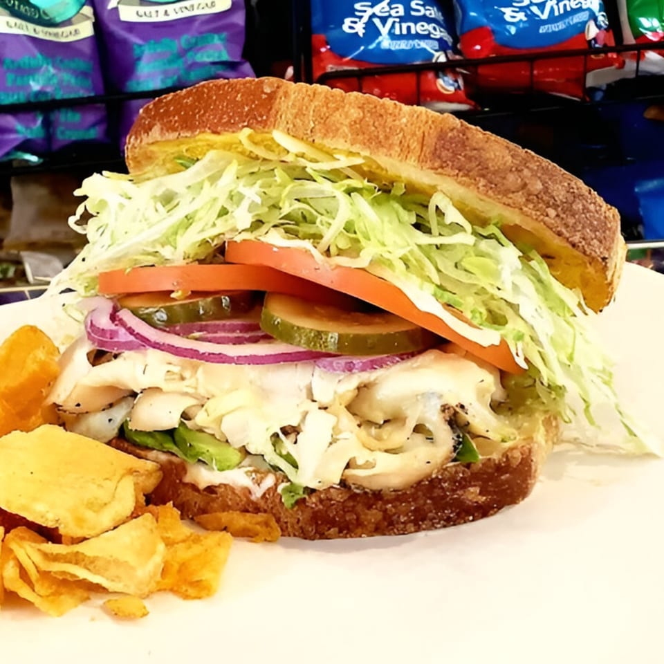 A Baba's Deli Sub with shredded lettuce, tomatoes, pickles, onions, and chips on the side.