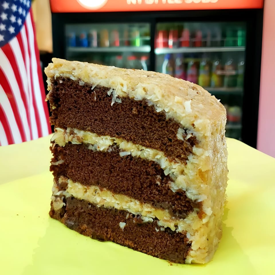 A slice of chocolate cake with coconut frosting on a yellow plate, with an American flag in the background.