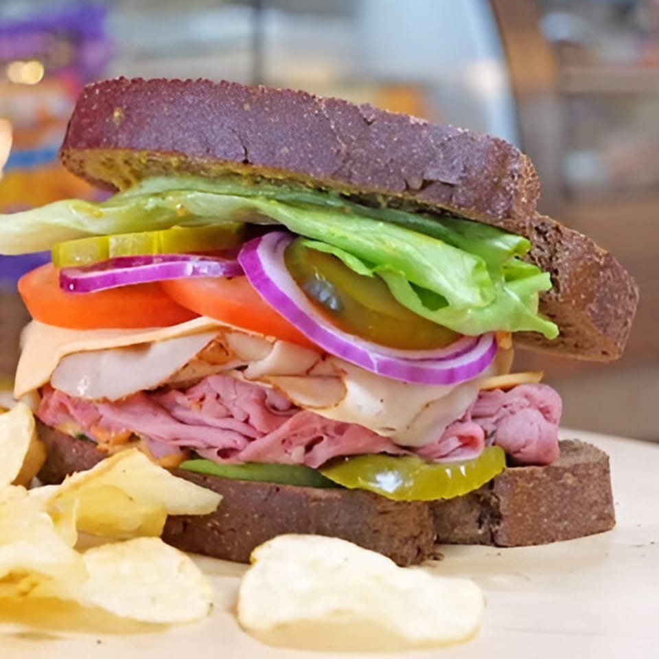 A Baba's Deli Sub with dark rye bread, piled high with meats, cheese, lettuce, tomato, onion, and pickles, served with potato chips.